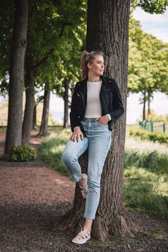 In einem Waldstück lehnt jemand an einem Baum. Er trägt eine schwarze Jacke, ein weißes Hemd, Jeans und ein Paar QLEAR Light Pink-Sneaker von ZAQQ, die für ihren hervorragenden Halt bekannt sind. Die Szene spielt sich vor grünen Bäumen und einem Pfad ab, durch dessen Blätter Sonnenlicht fällt.