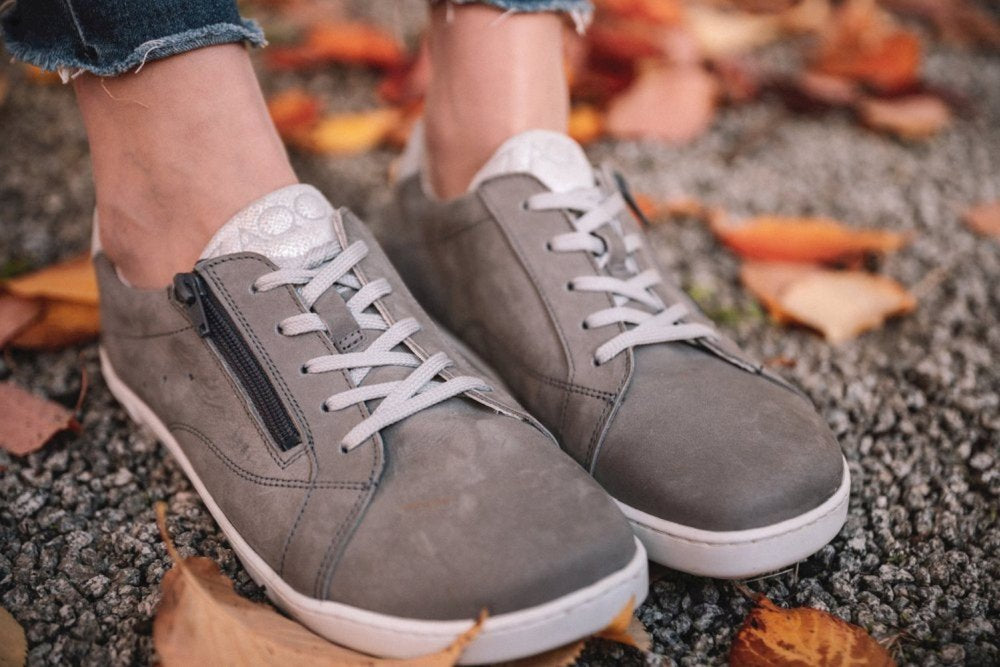 Eine Nahaufnahme zeigt jemanden, der auf einer Kiesfläche steht und die QOMFY Wide Grey Sneaker von ZAQQ trägt. Diese grauen Barfußschuhe aus Veloursleder sind mit weißen Schnürsenkeln und seitlichen Reißverschlüssen versehen und werden mit ausgefransten Jeans in Knöchellänge kombiniert. Gefallene Herbstblätter in Orange- und Rottönen umgeben ihre Füße und verstärken den natürlichen Komfort dieser Schuhe.