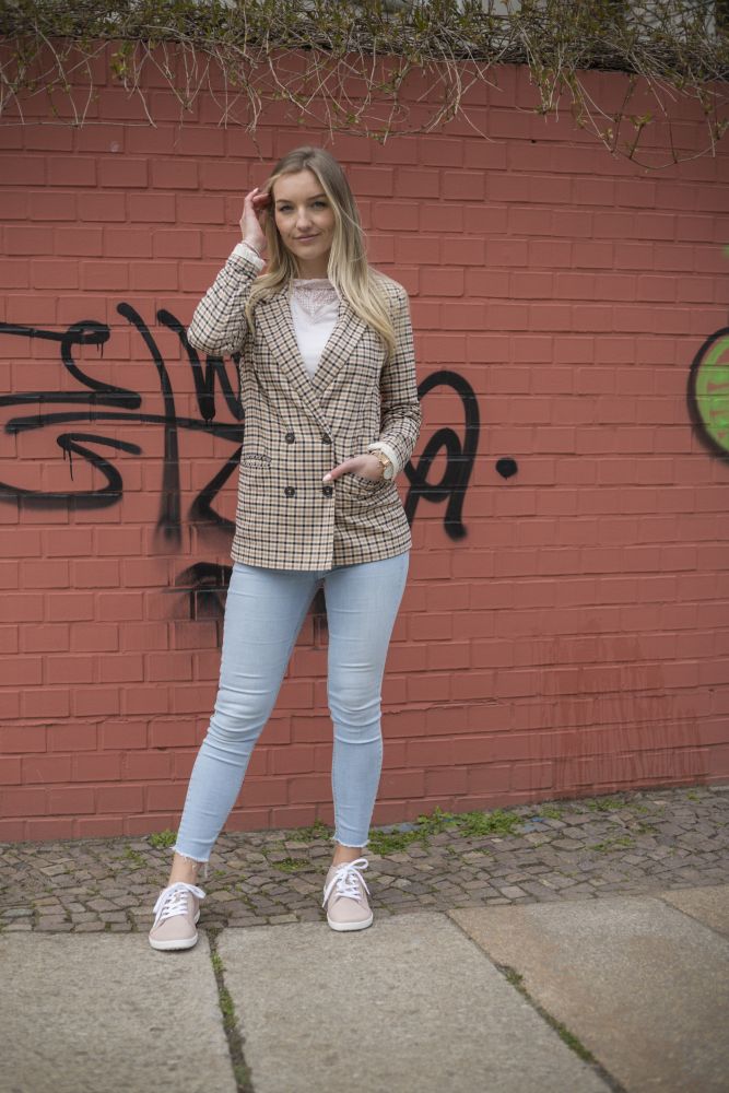 Eine Frau mit langen blonden Haaren steht vor einer mit Graffiti bedeckten Backsteinmauer. Sie trägt einen karierten Blazer, ein weißes Oberteil, hellblaue Jeans und beige Schuhe QOMFY Wide Rosa von ZAQQ. Sie berührt ihr Haar mit einer Hand und blickt selbstbewusst in die Kamera.