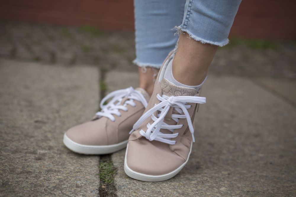 Eine Person trägt die QOMFY Wide Rosa Sneakers von ZAQQ aus hochwertigem Nappaleder in Hellrosa mit weißen Schnürsenkeln und Bluejeans mit ausgefranstem Saum und steht auf einem gepflasterten Platz.