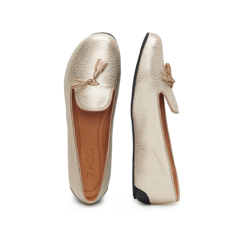 Die QOSMO Champagne Loafer von ZAQQ haben ein metallisches Gold-Finish mit einem schlichten, minimalistischen Design, das durch ein Schleifendetail auf der Oberseite ergänzt wird. Die schwarzen Sohlen aus hochwertigem Nappaleder bilden einen eleganten Kontrast zum Obermaterial, während die Barfuß-Loafer-Einlegesohlen den Markennamen ZAQQ in Braun tragen.