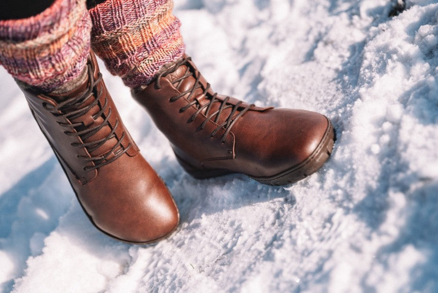 Eine Person, die ZAQQs QUINTIC Winter Vegan Nut Stiefel aus braunem Leder trägt, ergänzt durch bunt gestrickte Beinstulpen, steht im Schnee.