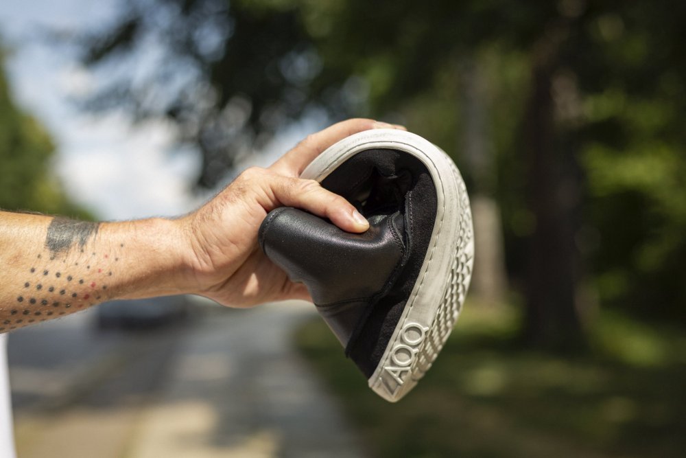 Eine Hand hält einen eleganten schwarzen QUITMAN-Sneaker von ZAQQ, der in der Mitte gebogen ist, um seine beeindruckende Flexibilität zu demonstrieren. Der Sneaker hat eine weiße Sohle mit dem Aufdruck „OOF“. Verschwommenes Grün und ein Bürgersteig bilden den Hintergrund und heben die natürliche Umgebung hervor.