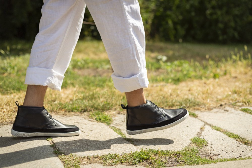 Eine Person in hochgekrempelten weißen Hosen und eleganten schwarzen QUITMAN-Sneakern von ZAQQ schreitet auf einem Steinpfad voran, im Hintergrund sind Gras und Grün zu sehen.