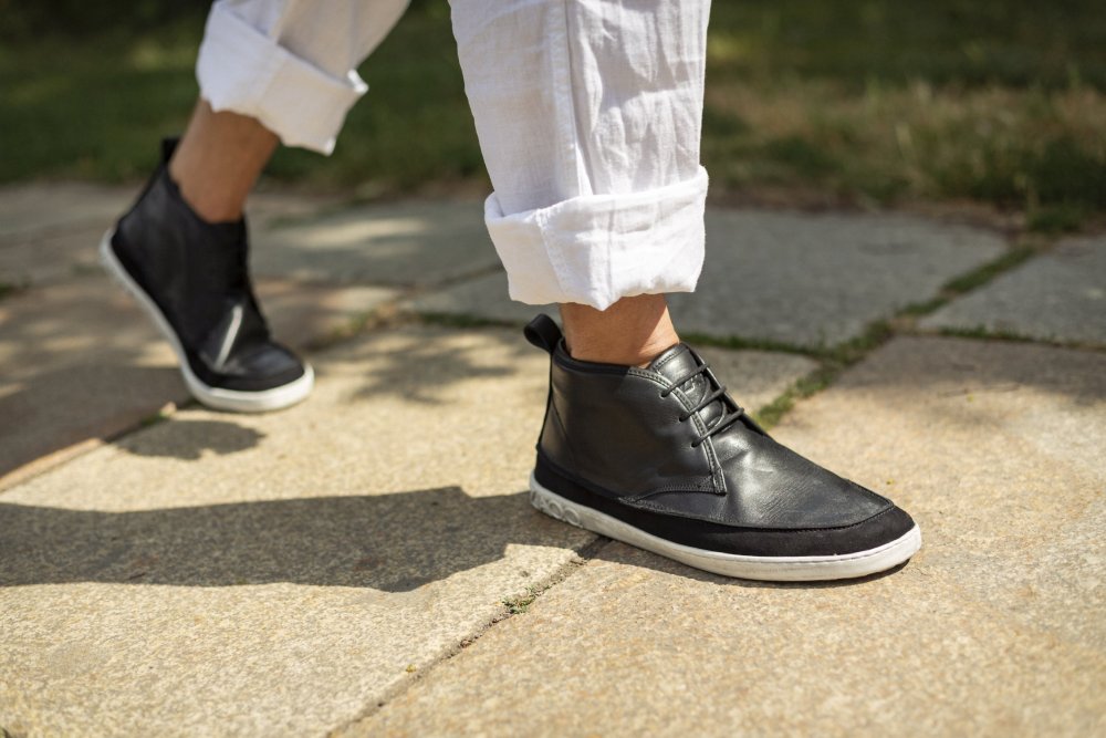 Eine Person in den stylischen QUITMAN Black Sneakers von ZAQQ und hochgekrempelten weißen Hosen läuft auf einem sonnenbeschienenen Steinpflaster, im Hintergrund ist Gras zu sehen.