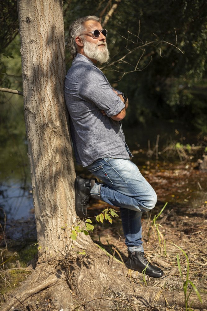 Ein bärtiger Mann mit Sonnenbrille lehnt in einem Waldstück an einem Baum. Er trägt ein blaues Hemd, Jeans und stylische schwarze ROQA-Stiefel von ZAQQ mit einem eleganten Reißverschluss. Er hat die Arme verschränkt und ein Bein angewinkelt. Das Sonnenlicht fällt durch die Bäume und wirft gesprenkelte Schatten auf den Boden und das nahe gelegene Wasser.