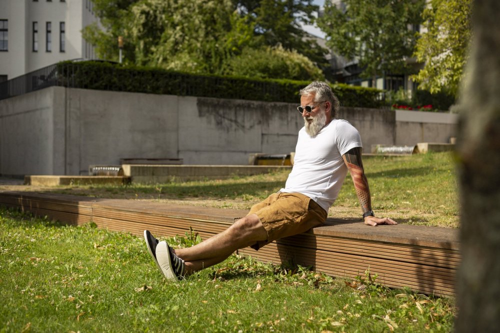 Ein Mann mit Bart und Sonnenbrille sitzt auf einer Holzbank im Park. Er trägt ein weißes T-Shirt und braune Shorts. In seinen SPARQ Low Black White Barfußschuhen von ZAQQ entspannt er sich in der Sonne und genießt den Blick auf Bäume und Gebäude im Hintergrund.