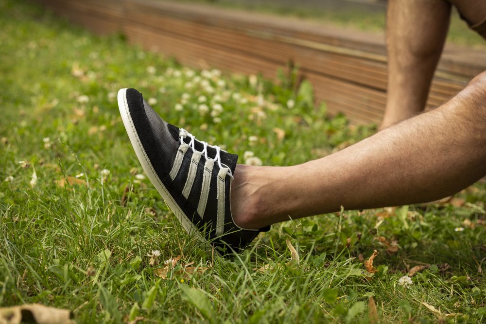 Eine Person sitzt mit ausgestreckten Beinen auf dem Gras und trägt die SPARQ Low Black White Sneaker von ZAQQ. Im Hintergrund sind eine Holzkonstruktion und weiteres Gras mit kleinen weißen Blumen zu sehen.