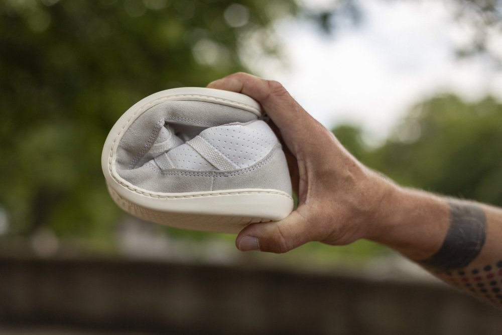 Eine Hand hält einen SPARQ Low White Sneaker von ZAQQ und biegt ihn mühelos in der Mitte, um seine Weichheit und seinen Komfort zu demonstrieren. Der helle Schuh mit der makellos weißen Sohle wird im Freien elegant vor einem verschwommenen grünen Hintergrund präsentiert.