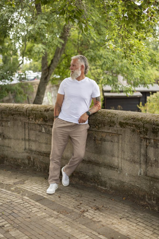 Ein Mann mit Bart, der ein weißes T-Shirt, beige Hosen und SPARQ Low White-Sneaker von ZAQQ trägt, lehnt an einem moosbedeckten Steingeländer. Er blickt in die Ferne in einer Parklandschaft, umgeben von üppigen grünen Bäumen. Seine Kleidung zeugt von mühelosem Stil und höchstem Komfort.