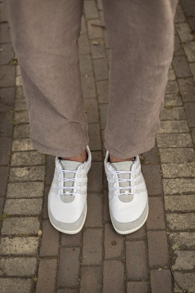 Eine Person in hellbraunen Hosen und bequemen SPARQ Low White Sneakers von ZAQQ steht auf einem mit Spitzen übersäten Weg und hebt das stylische Design und den Tragekomfort des Barfußschuhs hervor.