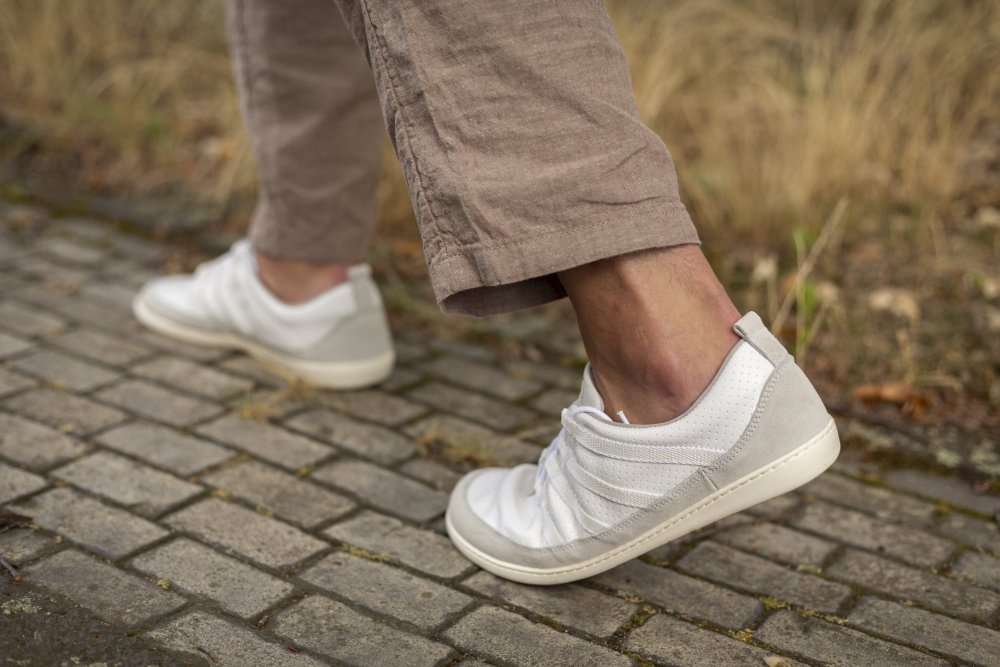 Eine Person schreitet in weißen SPARQ Low-Sneakers und einer braunen Hose einen Kopfsteinpflasterweg entlang, wobei ihre Schritte den Komfort der ZAQQ-Schuhe inmitten des trockenen Grases unterstreichen.