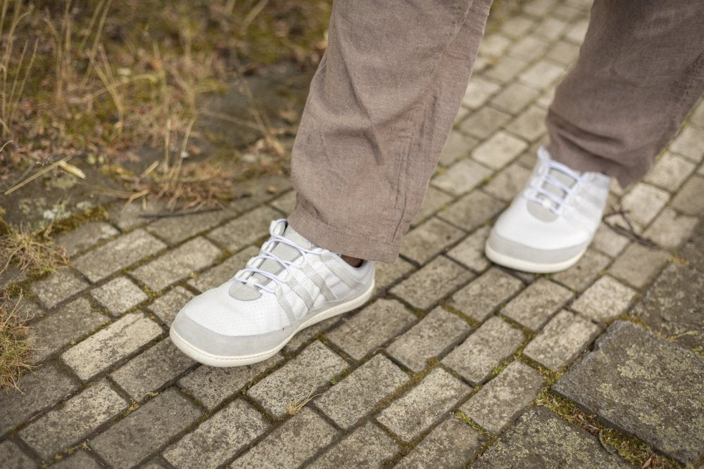 Eine Person in grauer Hose und SPARQ Low White Sneakers von ZAQQ läuft auf einem Kopfsteinpflasterweg. Gras und Moos schimmern durch die Steine und verstärken die Outdoor-Atmosphäre. Die Szene strahlt ein natürliches und lässiges Gefühl aus, wobei der Schwerpunkt auf Komfort liegt.