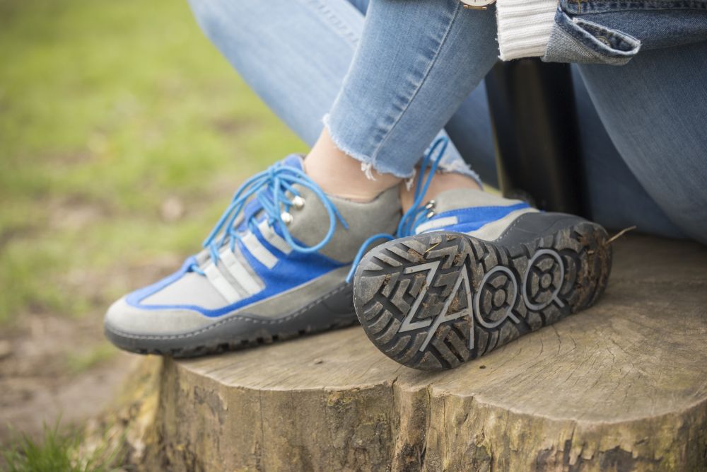 Eine Person trägt SQOUT Grey Blue Waterproof Sneakers von ZAQQ, die ein markantes Profilmuster aufweisen, und sitzt auf einem Baumstumpf. Diese Barfußschuhe sind sowohl atmungsaktiv als auch stylisch. Die Person trägt hellblaue Jeans und ein lässiges Oberteil und genießt die Natur auf einer Rasenfläche.