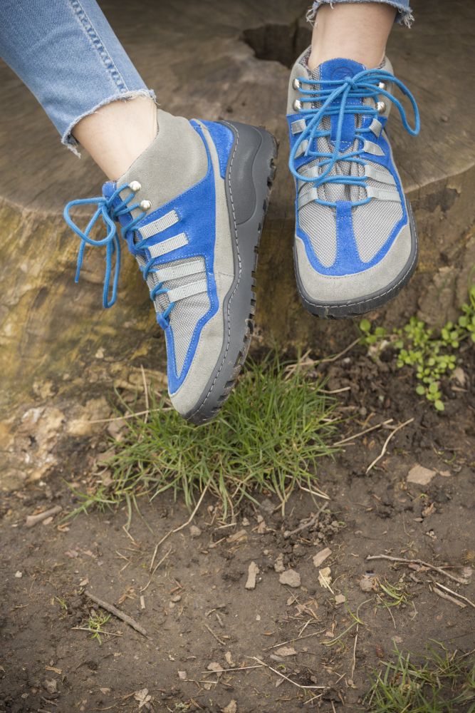 Ein Paar SQOUT Grey Blue Waterproof Wanderschuhe von ZAQQ, bekannt für ihre wasserdichte Eigenschaft, ruht auf einem großen Baumstumpf. Die hellblauen Schnürsenkel ergänzen die schwarze Sohle. Darunter befindet sich eine Mischung aus Erde, kleinen grünen Pflanzen und Gras.