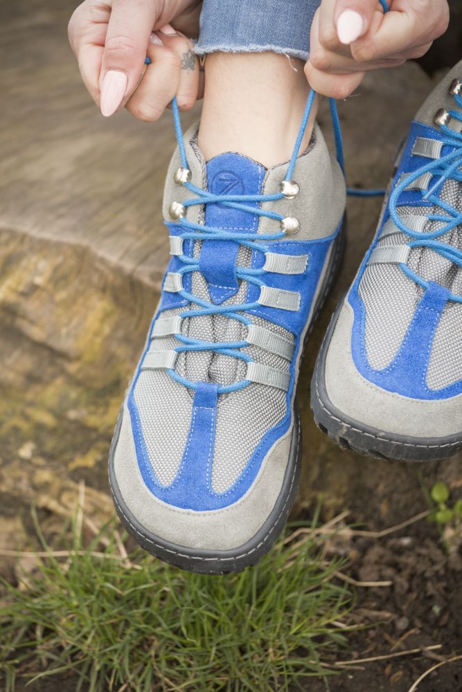 Eine Nahaufnahme von jemandem, der auf einem Baumstumpf sitzt und die Schnürsenkel der ZAQQ SQOUT grau-blauen wasserdichten Wanderschuhe bindet. Die Person trägt Jeans und in der Nähe des Stumpfes ist Gras zu sehen, was das atmungsaktive Design hervorhebt, das ideal für Outdoor-Abenteuer ist.