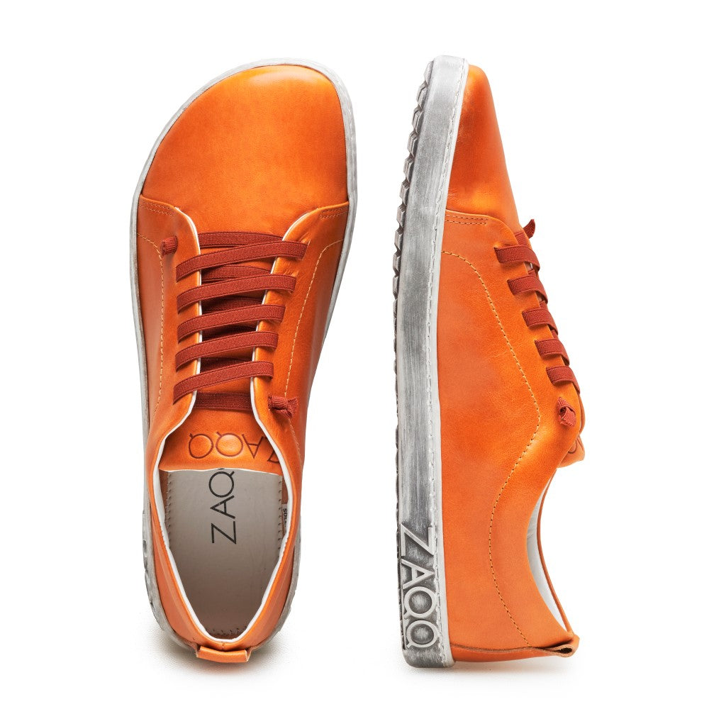 Wir präsentieren den STIQ Sierra von ZAQQ: Ein auffälliges Paar leuchtend orangefarbener Sneaker aus Nappaleder mit auffälligen braunen Schnürsenkeln und grauen Sohlen. Der linke Sneaker wird elegant von der Seite präsentiert, während der rechte von oben betrachtet wird und das markante Zaoo-Logo auf der Innensohle und entlang der Sohlenkante zeigt. Diese schicken Barfußschuhe vereinen mühelos Komfort und Stil.