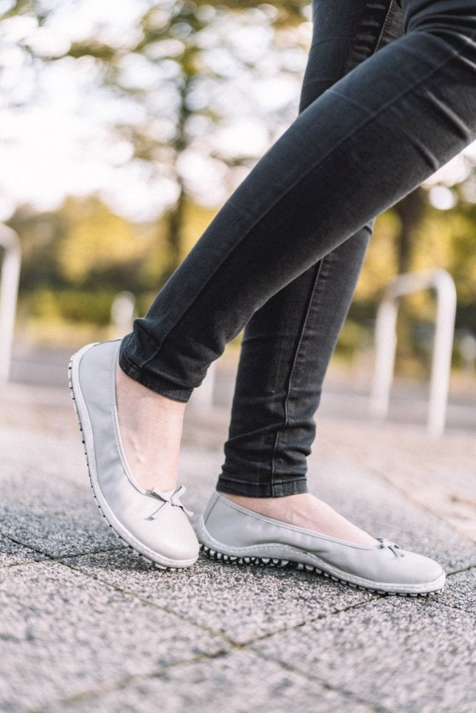 Eine Person in stylischen SUQAR Grey Flats von ZAQQ steht im Freien auf einer strukturierten Steinoberfläche. Sie trägt schwarze Hosen, der Hintergrund zeigt verschwommenes Grün und weiße Geländer, wodurch eine harmonische Mischung aus Eleganz und Natur entsteht.