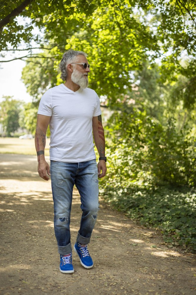 Ein Mann mit weißem Bart und Sonnenbrille schlendert einen Feldweg im Park entlang. Seine wasserdichten TERRAQ Azure-Schuhe von ZAQQ ergänzen seinen lässigen Look aus weißem T-Shirt, Blue Jeans und blauen Sneakers. Grüne Bäume und Sonnenlicht zieren den Hintergrund.