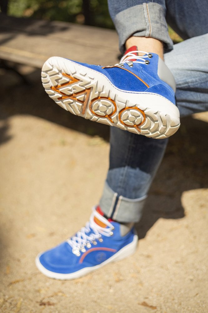 Eine Person sitzt mit gekreuzten Beinen auf einer Bank und präsentiert die blauen TERRAQ Azure Waterproof Sneaker von ZAQQ, sorgfältig aus Veloursleder mit weißer Sohle gefertigt. Die Jeans ist hochgekrempelt und gibt den Blick auf gestreifte Socken frei. Diese Sneaker haben orangefarbene Akzente und ein einzigartiges Profil, perfekt für jedes Outdoor-Abenteuer. Sonnenlicht wirft weiche Schatten.