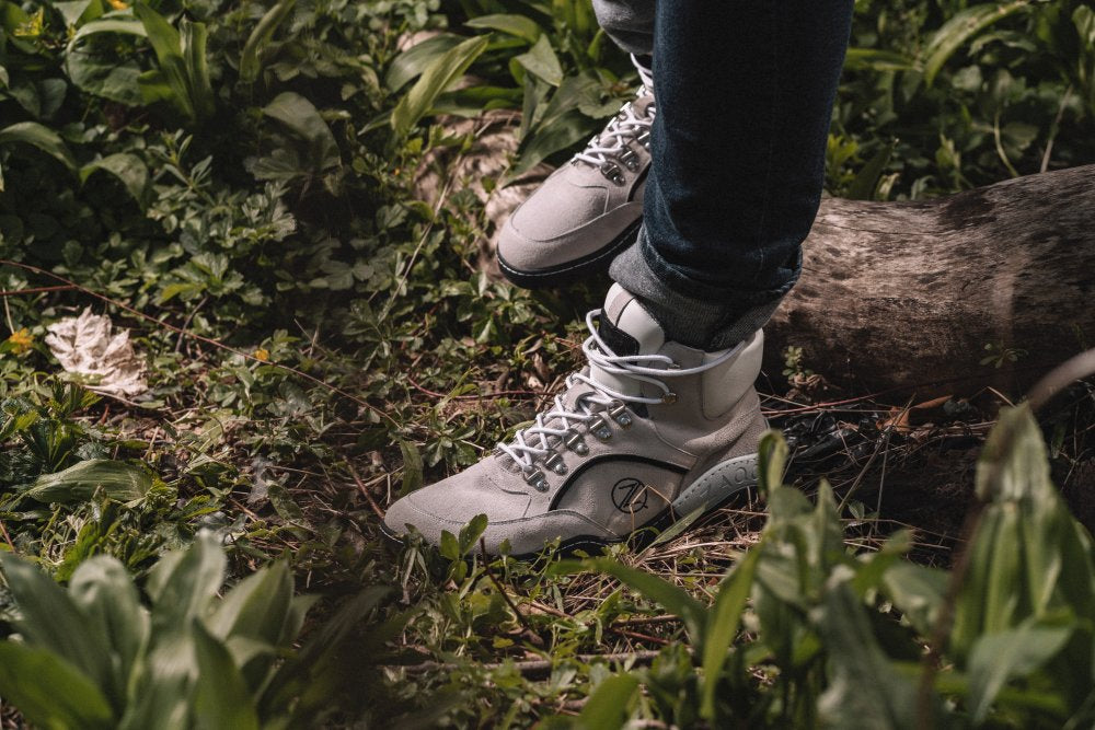 Eine Person, die die wasserdichten High-Top-Sneaker ZAQQ TERRAQ Beige aus Veloursleder trägt, steht auf grasbewachsenem Boden inmitten von Blattpflanzen, in der Nähe liegt ein umgestürzter Baumstamm.