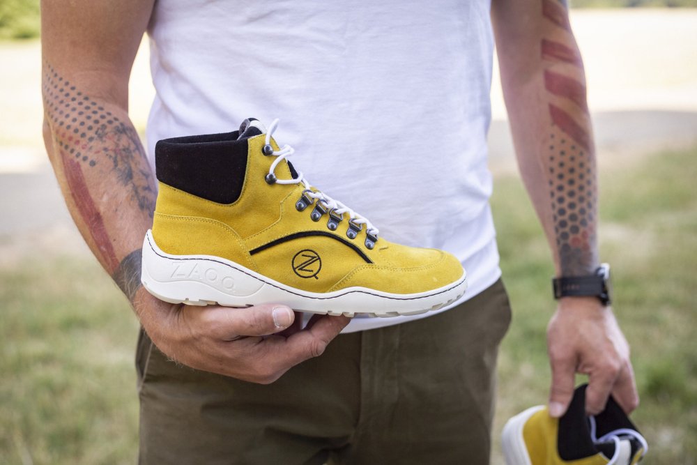 Eine Person hält den TERRAQ Yellow Waterproof Sneaker von ZAQQ in der Hand, ein leuchtend gelbes High-Top-Design aus Wildleder mit weißer Sohle und auffälligen schwarzen Akzenten. Der Schuh ist mit Schnürsenkeln ausgestattet und trägt das Logo deutlich sichtbar auf der Sohle. Die Person hat bunte Tattoos auf den Unterarmen und trägt ein weißes Hemd und olivfarbene Hosen.