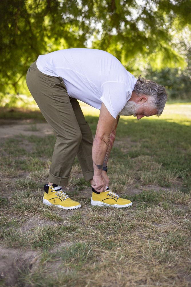 Ein älterer Mann mit grauem Haar und Bart bückt sich, um seine leuchtend gelben TERRAQ Yellow Waterproof-Sneaker von ZAQQ zu binden. Er trägt ein weißes T-Shirt und eine grüne Hose und steht auf einer Wiese mit Bäumen im Hintergrund.