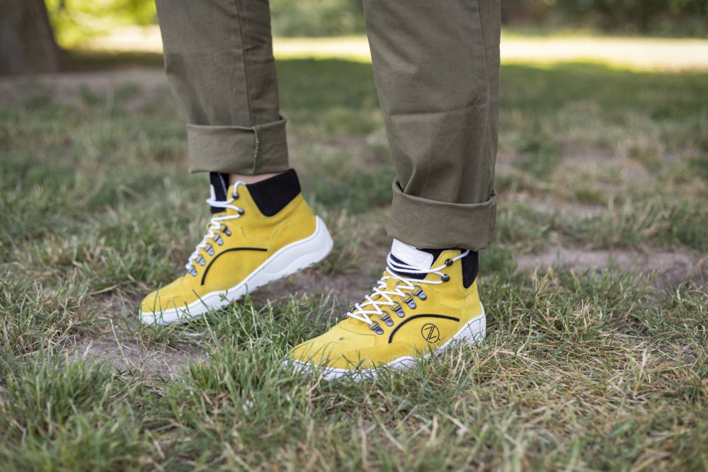 Eine Person, die TERRAQ Yellow Waterproof High-Top-Sneakers von ZAQQ mit weißen Sohlen und schwarzen Akzenten trägt, steht auf Gras. Ihre Khakihose ist an den Beinenden hochgekrempelt und zeigt die perfekte Kombination aus Stil und Funktionalität für jedes Abenteuer.