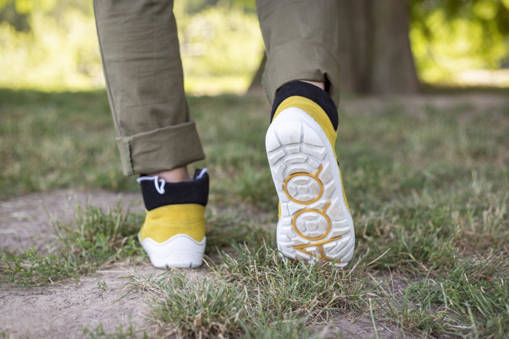 Eine Person trägt ZAQQs TERRAQ Yellow Waterproof-Sneaker mit weißer Sohle und olivgrüne Hose und spaziert über eine Wiese. Die Sohle eines Sneakers zeigt die Nummer 400 im Profildesign, was seinen Stil und seine Zweckmäßigkeit für jeden Ausflug ins Freie unterstreicht.