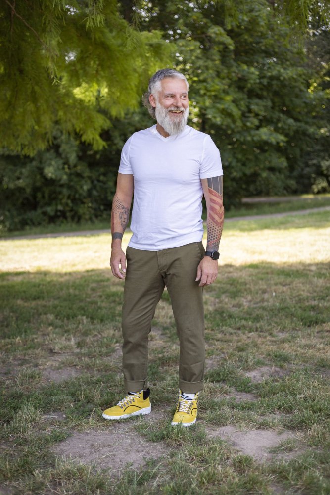 Ein lächelnder Mann mit grauem Bart und Tattoos steht draußen auf einer Wiese. Er trägt ein weißes T-Shirt, olivgrüne Hosen und leuchtend gelbe wasserdichte Turnschuhe aus der TERRAQ-Kollektion von ZAQQ. Im Hintergrund sind Bäume zu sehen.