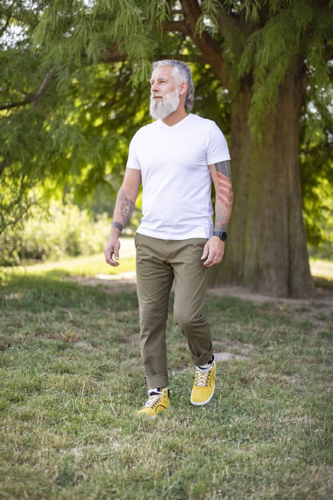 Ein bärtiger Mann mit Tattoos schlendert durch einen grasbewachsenen Park. Er trägt ein weißes T-Shirt, olivgrüne Hosen und leuchtend gelbe wasserdichte ZAQQ TERRAQ Sneakers. Ein großer Baum mit üppigem grünem Laub bietet die perfekte Kulisse für sein entspanntes Outdoor-Abenteuer.