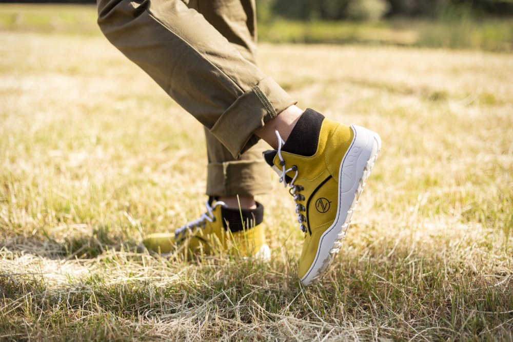 Eine Person, die TERRAQ Yellow Waterproof Sneakers von ZAQQ trägt, mit hohen Oberteilen, weißen Sohlen und dunklen Schnürsenkeln, steht auf trockenem Gras. Sie trägt hochgekrempelte Khakihosen vor einer sonnigen Außenkulisse, die diese robusten Outdoorschuhe perfekt zur Geltung bringen.