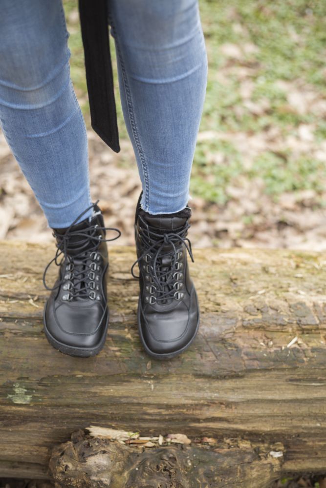 Eine Person, die die wasserdichten TREQ Black Winter-Stiefel von ZAQQ und Blue Jeans trägt, steht auf einem großen umgestürzten Baumstamm, umgeben von einem mit Blättern und Gras bedeckten Boden.