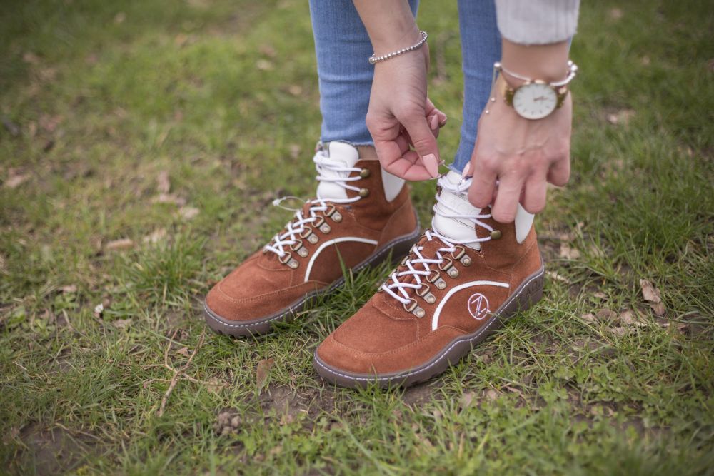 Eine Person in Bluejeans und weißem Pullover bindet die Schnürsenkel ihrer wasserdichten braunen TREQ Cognac Wanderschuhe von ZAQQ und steht auf einer Wiese. Sie trägt eine Armbanduhr und ein Armband.