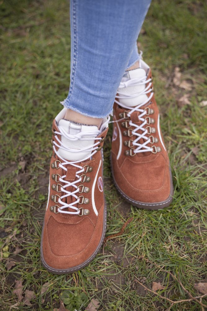 Nahaufnahme einer Person, die ZAQQs wasserdichte Stiefel TREQ Cognac mit weißen Schnürsenkeln trägt und auf Gras steht. Die Person trägt hellblaue Jeans und der Boden besteht aus einer Mischung aus grünem Gras und Schmutzflecken.