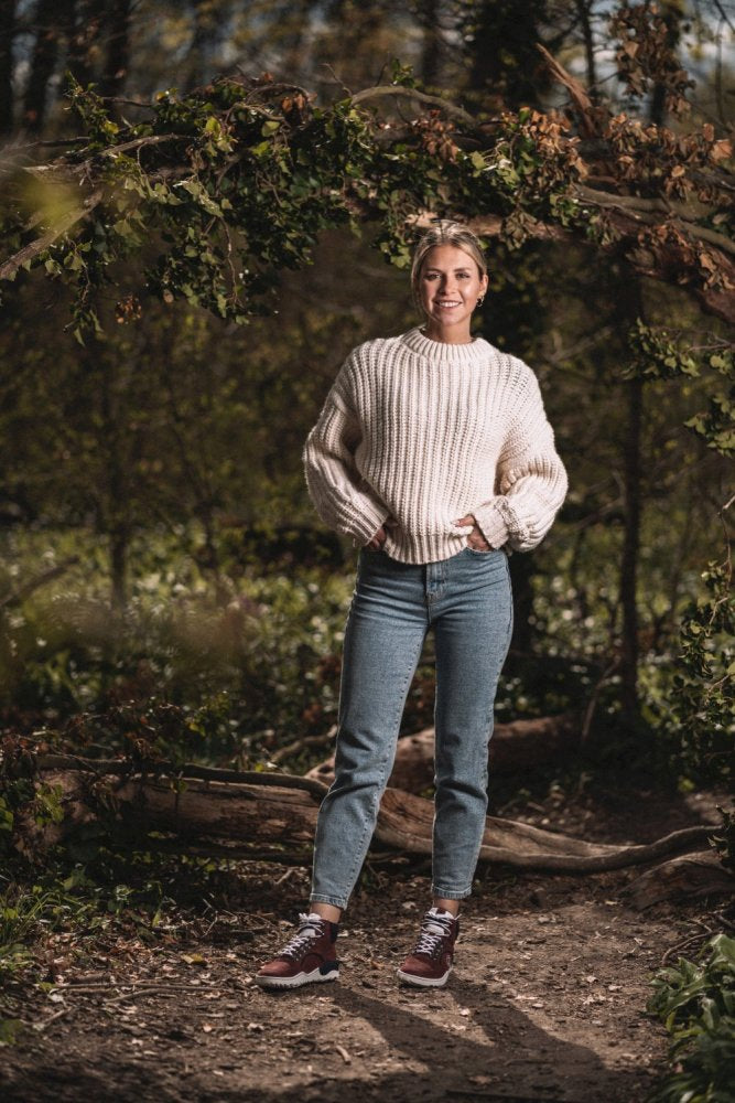 Eine Person steht auf einem Waldweg und trägt einen cremefarbenen Pullover, Jeans und die TREQ Red Waterproof-Sneakers von ZAQQ. Sie ist von Grün umgeben und Sonnenlicht fällt durch die Bäume darüber und schafft eine ruhige Atmosphäre.