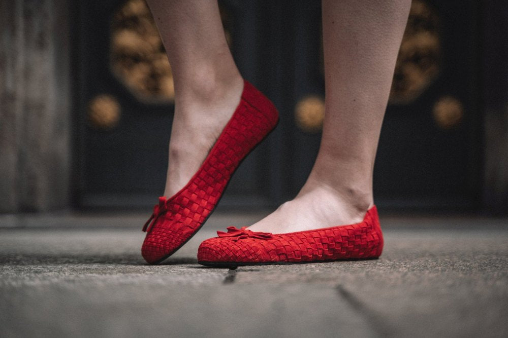 Eine Person, die die TWIST Red Ballerinas von ZAQQ mit gewebter Textur trägt, steht auf einer rauen Steinoberfläche. Diese mit kleinen Schleifen verzierten Velourlederschuhe ziehen die Aufmerksamkeit auf sich, da ein Fuß leicht angehoben ist. Der unscharfe Hintergrund unterstreicht den Stil und die Eleganz dieser von Barfuß-Ballerinas inspirierten Ballerinas.