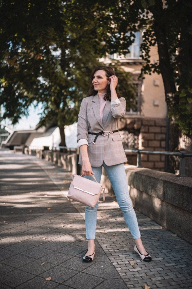 Eine Frau steht auf einem von Bäumen gesäumten Gehweg. Sie trägt einen karierten Blazer, hellblaue Jeans und TWIST Varnish Nude Flats von ZAQQ mit Gummisohle. Sie hält eine hellrosa Handtasche in der Hand, ordnet mit einer Hand ihr Haar und genießt den sonnigen Tag.