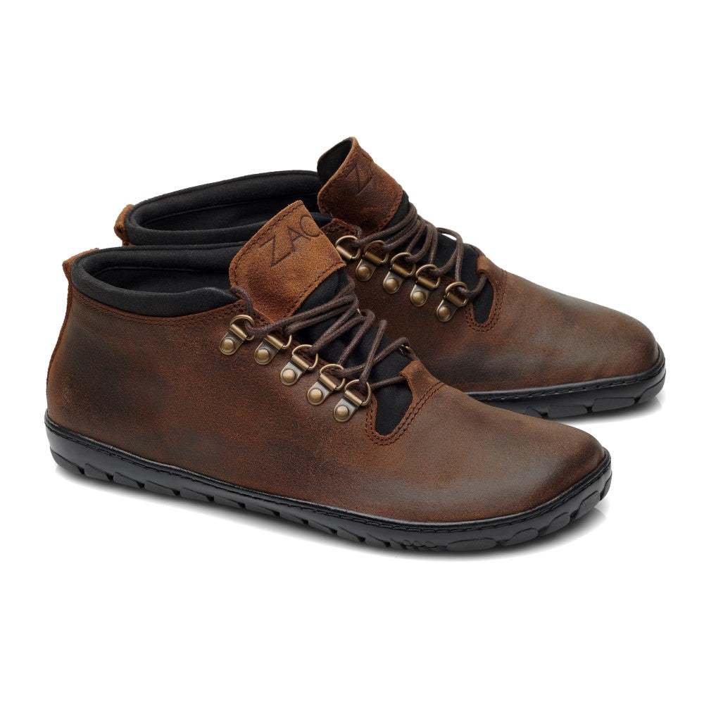 Der EXPEQ Mid Brown Waterproof von ZAQQ ist ein Paar brauner Leder-High-Top-Schuhe, der als Outdoorschuh konzipiert ist und über eine schwarze Sohle und messingfarbene Ösen verfügt. Diese Schuhe haben schwarze Schnürsenkel, eine leicht raue Oberfläche und sind wasserdicht. Das Markenlogo ist elegant auf der Zunge geprägt.