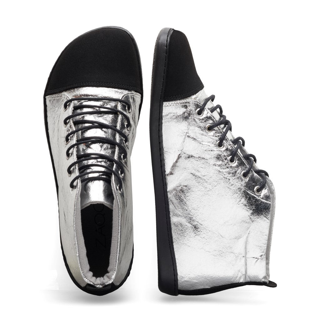 Diese QIVO Mid Pinatex-Sneakers präsentieren die Marke ZAQQ und zeichnen sich durch ein veganes High-Top-Design in Silber mit schwarzen Sohlen und Schnürsenkeln aus. Ein Schuh steht aufrecht und betont seine elegante Seite, während der andere flach liegt und die sauber gebundenen Schnürsenkel vorne zeigt, was sowohl Stil als auch außergewöhnlichen Komfort gewährleistet.