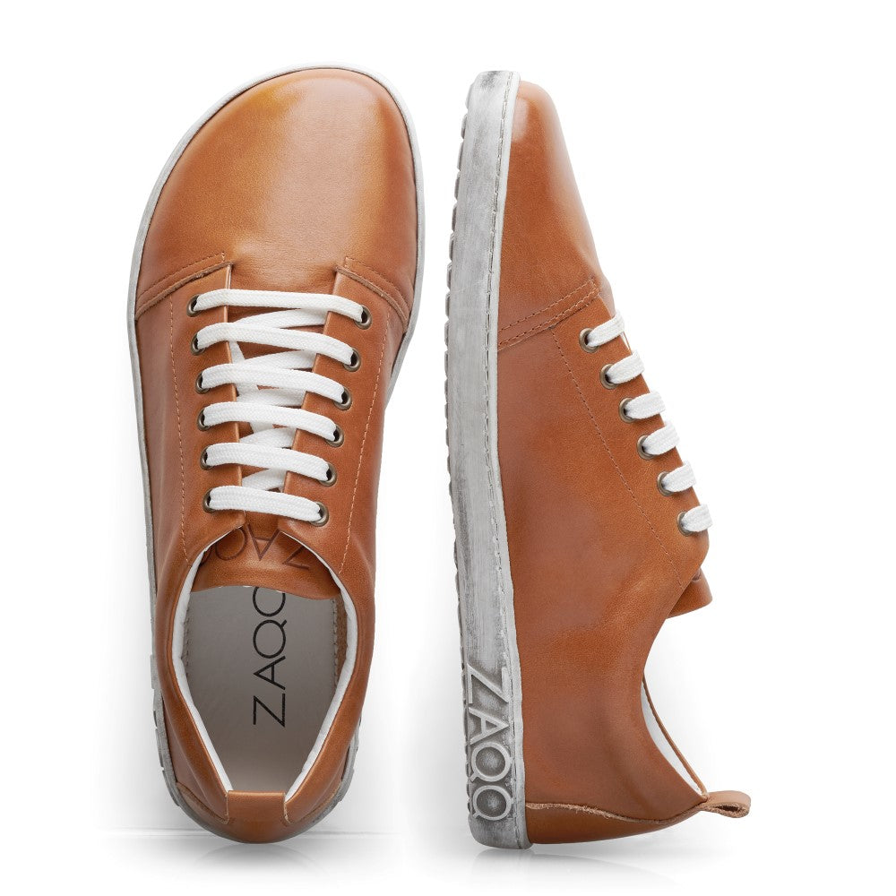 Ein Paar stylische TAQQ Nappa Brown-Sneaker von ZAQQ mit einer eleganten Außenseite aus braunem Nappaleder und weißen Schnürsenkeln. Ein Schuh steht aufrecht, während der andere auf der Seite liegt und die charakteristische flexible Barfußsohle der Marke hervorhebt. Diese Barfuß-Sneaker verbinden nahtlos lässige Eleganz mit unvergleichlichem Komfort.